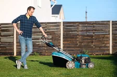 Gardening in the backyard. Young man in a plaid shirt with a push lawn mower on the green grassの写真素材