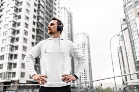Take a break at workout. Close-up portrait of young man in sports clothing with headphones stand while exercising outsideの写真素材