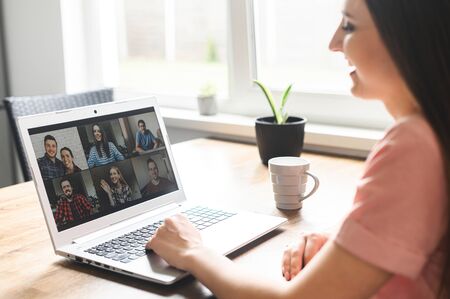 Communication on distance via video, video call, zoom. A young attractive woman using app on laptop for meeting with friends. She looks at webcam and smiles. Side viewの写真素材