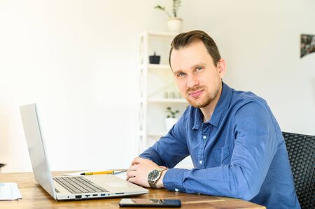 Work at home. A young handsome man is formal shirt is using laptop for remote work, he looks at the...