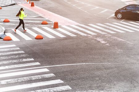 Morning jog during quarantine. Young woman in sportswear with a medical mask on the face runs on the zebra crossing, she is listening music in headphonesの写真素材