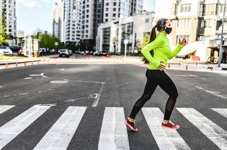 Morning jog during quarantine. Young woman in sportswear with a medical mask on the face runs on the road among residential skyscrapers, she is listening music in headphonesの写真素材