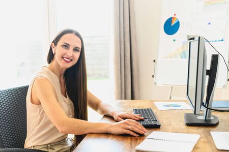 Work at office. A young happy woman is using pc for office work, she sits at the table and looks to the camera, flip chart on backgroundの写真素材
