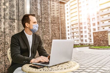 Epidemia in the city. A young businessman with a protective medical mask on the face is working with laptop on the outdoor summer terrace cafe in the downtownの写真素材