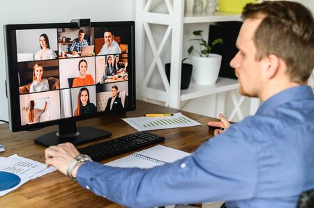 Virtual conference with employees. A young man in formal shirt using pc for video call, he has video meeting with several people together. Back view. Remote work concept.の写真素材
