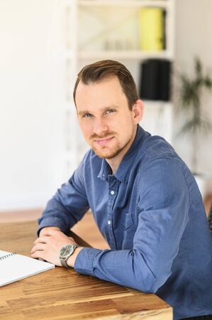 Office work. Portrait of young handsome man in smart casual shirt, he sits at the table and looks at camera with a smile. Vertical photoの写真素材