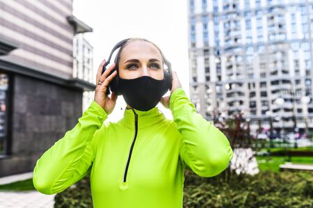 Doing sports on quarantine. Close-up portrait of a young woman in neon green sportswear with a headphones on her head in the urban landscapeの写真素材
