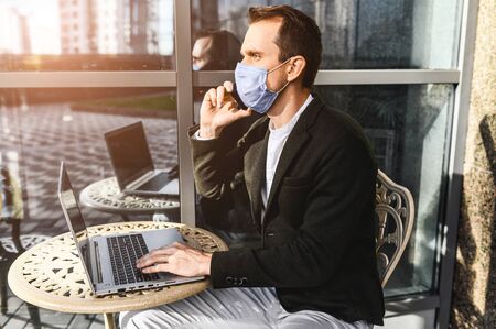 A young man in a medical mask using laptop for work outdoors. He sits on the summer terrace at the table and talking on the phone. Quarantine coronavirus, pandemia covidの写真素材