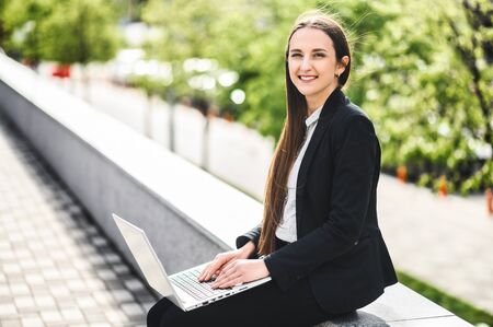 Portrait of young woman in smart casual wear with a laptop outdoors. She looks at camera and smiles. Remote workの写真素材