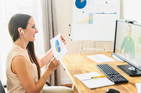 Video call, video meeting. A young woman is discussing some business tasks with a man via video, she sits at office space and showing paper with a graphs at webcamの写真素材