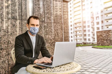 Epidemia in the city. A young businessman with a protective medical mask on the face is working with laptop on the outdoor summer terrace cafeの写真素材