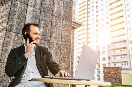 A young businessman in smart casual wear is working with laptop on the outdoor summer terrace cafe, he is sitting at the table and talking on the phoneの写真素材