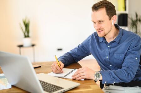 Online learning, webinars. A guy in smart casual clothes is watching video lectures on the laptop and doing notes in notebookの写真素材