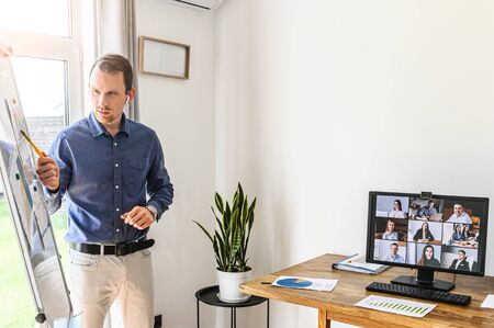 Online presentation, webinar, online meeting. A young man speaks to the online audience via video call, video connection. He stands near flip chart, pc screen with online viewers in front of himの写真素材