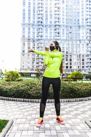 A young woman in trendy sportswear with a black medical mask on the face is doing warming up before workout outdoors. Sports training during quarantine. Vertical photoの写真素材