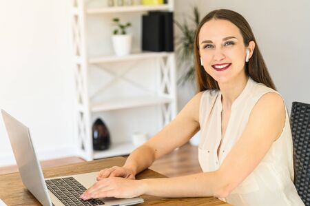 Home office. A young attractive woman is using laptop for remote work, she sits at the table, looks at camera and smilesの写真素材
