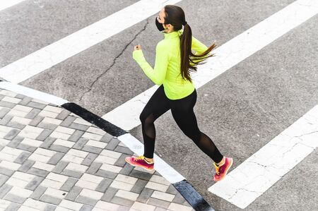 Sports training during quarantine. Full length young woman is running in medical mask on the roadの写真素材
