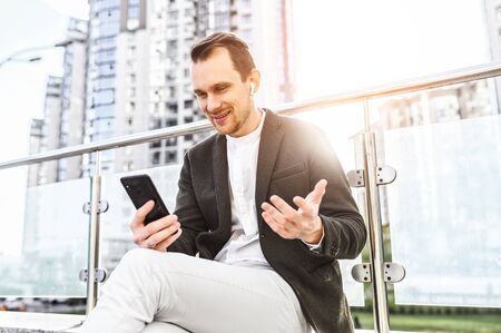 Video communication concept. A young businessnman is using phone for video call outdoors, the guy is talking via video and smiles, skyscrapers on background.の写真素材
