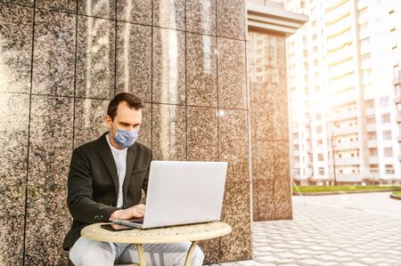 The guy uses a laptop to work outdoors during the coronavirus epidemic. He is sitting at the table on summer terrace of cafe among the skyscrapers with a medical mask on his face.の写真素材