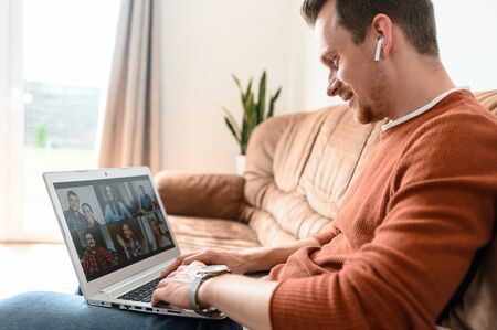 Video call in zoom. A guy is communicated with friends white staying at home. He sits on the sofa and using laptop for video chattingの写真素材