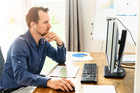 Work at office. A young man is using pc for office work, she sits at the table and concentrated looks to the screen, flip chart on backgroundの写真素材