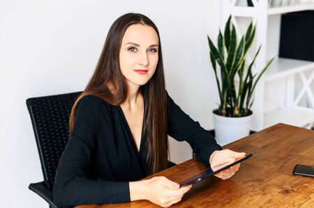 Beautiful woman in black shirt with a digital tablet in hands is sitting at the table in office and looks at cameraの写真素材