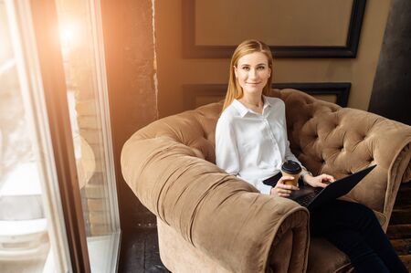 Office work in the loft style. Portrait of an attractive young girl in a formal white shirt with a laptop and coffee sitting in an armchair and looking at the cameraの写真素材