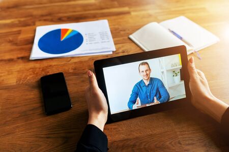 Video call, zoom. Webcam view of a young man in formal shirt on the tablet screen in female hands in the office, notebook and working papers on the table nearの写真素材