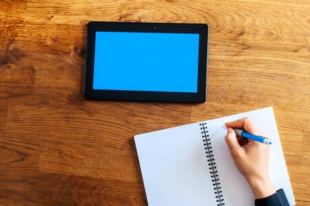 Top view of female hand is writing abstract by pen in a notebook, a digital tablet on a wooden table near. A screen of tablet is blank, copy space for text. Online learning conceptの写真素材