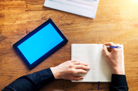 Top view male hands are writing by pen in notebook. A digital tablet with blank screen and working papers are near on the wooden table. Online learning concept. Flat layの写真素材