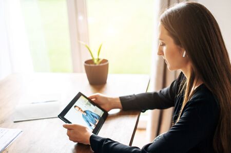 Video meeting. A young woman with digital tablet in smart casual wear is talking via video call with a man in office. Side viewの写真素材