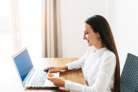 A woman using laptop for online shopping, online banking. She sits at the table with a credit card in hand and looks at the screen with a smileの写真素材