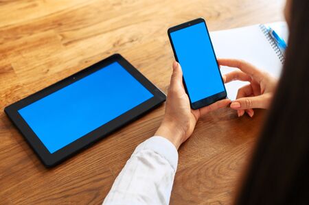 A young woman is using gadgets for work in office. Back view of phone with blank screen in female hands, a digital tablet is near on the table. Copy spaces for the text, mockupの写真素材