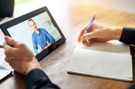 Close-up male hand is wting by pen in notebook, a gital tablet with a online tutor, online teacher on the screen in other hand. Online classes, webinarsの写真素材