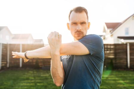Attractive bearded guy is doing stretching exercises outdoors, he is warming up before workout in the backyardの写真素材