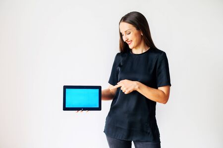 A young cheerful woman in black wear is holding a blank digital tablet in hand, pointing finger of other hand at the empty screen, isolated on white background. Copy space for textの写真素材