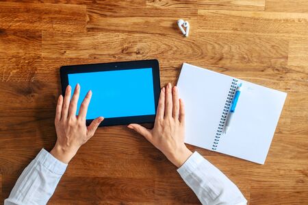Top view female hands in formal white shirt with a digital tablet with a blank screen on a wooden table, a notebook with a pen for notes is nearの写真素材
