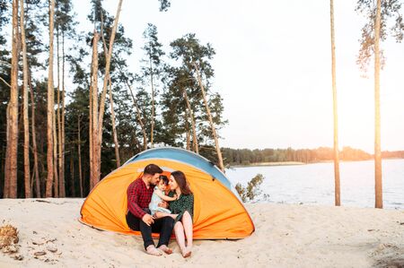 Young family spend time together outdoors. Beautiful mother, handsome father and little cute daughter spend active weekend in a camp, they are sitting in a tent surrounded a forestの写真素材