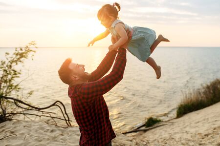 Fathers day. Young handsome dad with a beard spends time with his cute daughter outdoors. Happy father are throws up a laughed girl, lake on backgroundの写真素材