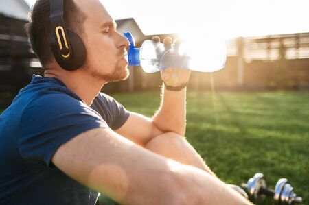 The guy quenches thirst during an outdoor workout. He sits with a headphones and drinks water from a sports bottleの写真素材