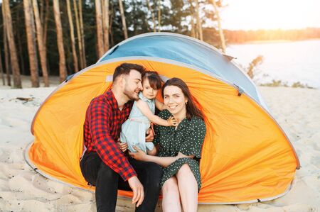 Young family spend time together outdoors. Beautiful mother, handsome father and little cute daughter spend active weekend in a camp, they are sitting in a tent surrounded a forestの写真素材