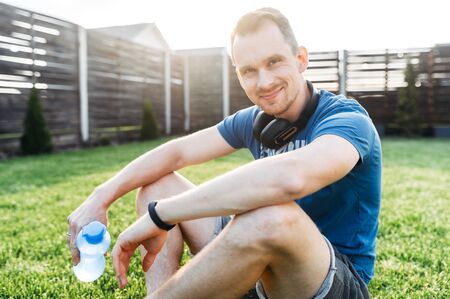 Healthy lifestyle. Portrait of a guy in sports wear with a bottle of water, fitness tracker on his wrist. He is taking a break between sets while doing outdoor workout, looking at camera and smilesの写真素材