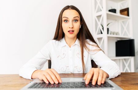 Webcam shot of a young surprised woman in white shirt, she is typing on the laptop keyboard and looks at the screen with big eyes. Unexpected newsの写真素材