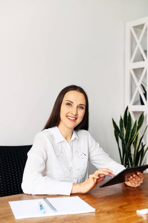 A young cheerful beautiful woman in formal white shirt sits at the table in office with a tablet in hands. She smiles and looks at cameraの写真素材