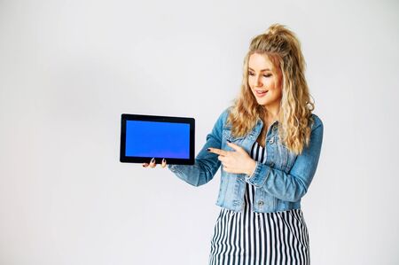 Look at this. Beautiful young woman with a tablet computer in hands stands on a white background. Woman points finger to blank tablet screen, empty space for inserting pictures, textの写真素材
