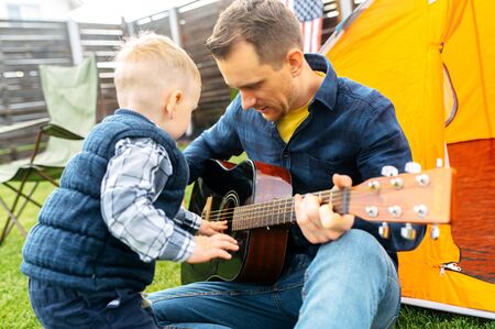 Father and son spend time in the backyard. Daddy plays guitar, toddler son plays nearの写真素材