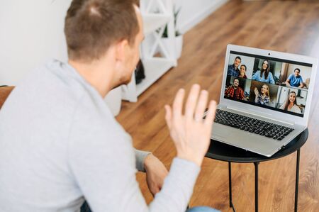 A guy waving hello into webcam and smiles. A guy is using app on the laptop for video chat with a several friends at same timeの写真素材