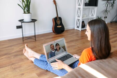 A young woman is talking online with her parents via video call on the laptop in a cozy homely atmosphere. Communication with a family on the distanceの写真素材