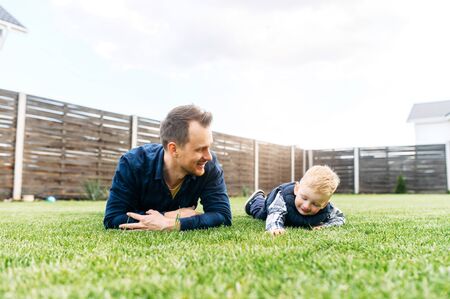 Father and little son are lying on the green lawn and laugh. Dad and toddler play in the backyardの写真素材