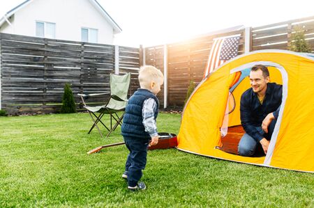 Father and son play camp in the backyard. Young father and his little toddler boy are playing together in an orange tent outdoors. Folding chair, guitar are near on the lawnの写真素材